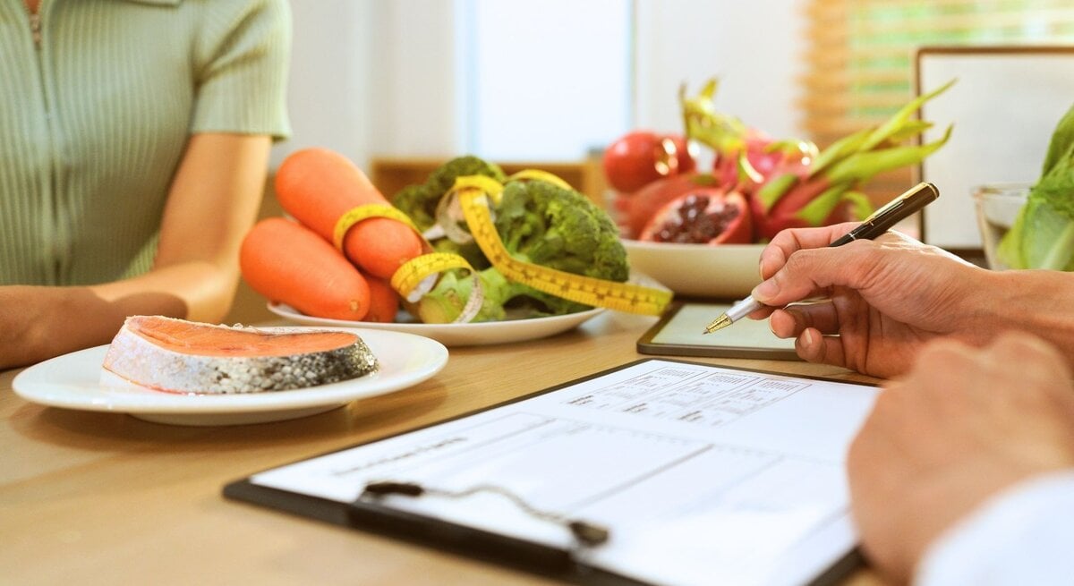 Zwei Personen bei einer Ernährungsberatung: links sitzt eine Person in einem grünen Oberteil, rechts hält eine Hand einen Stift über einem Klemmbrett mit Ernährungsplan. Auf dem Tisch liegen ein Teller mit Lachsfilet sowie Schalen mit Karotten, Brokkoli und aufgeschnittenem Granatapfel; ein Maßband ist um das Gemüse gelegt, Tageslicht fällt durch das Fenster.