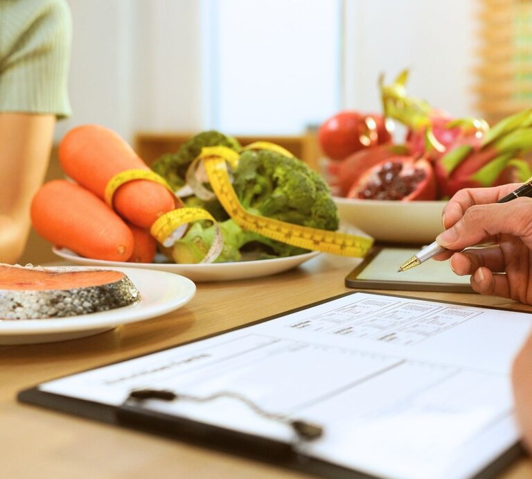 Zwei Personen bei einer Ernährungsberatung: links sitzt eine Person in einem grünen Oberteil, rechts hält eine Hand einen Stift über einem Klemmbrett mit Ernährungsplan. Auf dem Tisch liegen ein Teller mit Lachsfilet sowie Schalen mit Karotten, Brokkoli und aufgeschnittenem Granatapfel; ein Maßband ist um das Gemüse gelegt, Tageslicht fällt durch das Fenster. | © Thidarat kwangten via Canva.com 
