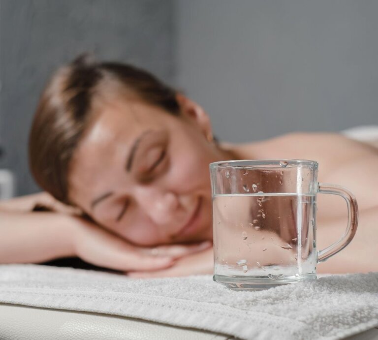 Eine Frau liegt antspannt auf einer Massageliege. Im Vordergrund ein Glas Wasser | © Alamy/ Nina Hilitukha via Canva.com
