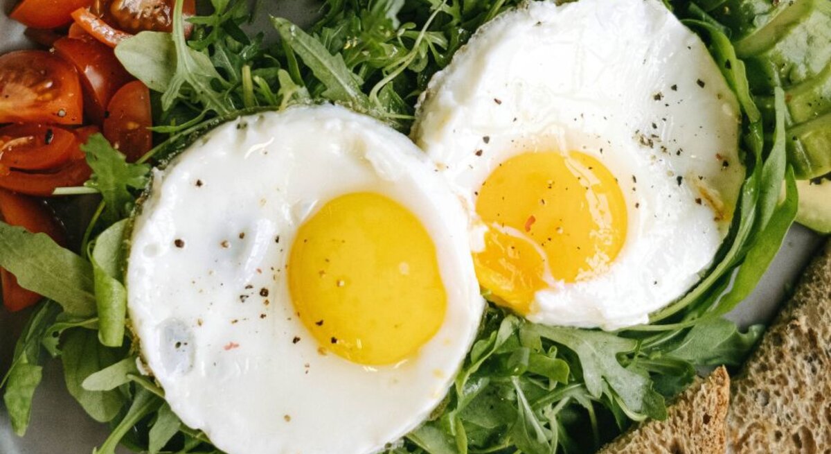 Zwei Spiegeleier mit leuchtend gelbem Eigelb, angerichtet auf frischem Rucola. Dazu gibt es frische, saftige Tomatenstücke, Avocadoscheiben und eine Scheibe Vollkornbrot, serviert auf einem grauen Teller.