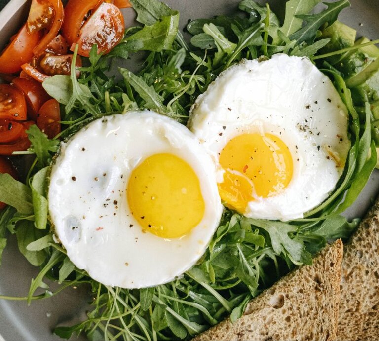 Zwei Spiegeleier mit leuchtend gelbem Eigelb, angerichtet auf frischem Rucola. Dazu gibt es frische, saftige Tomatenstücke, Avocadoscheiben und eine Scheibe Vollkornbrot, serviert auf einem grauen Teller. | © Pexels/ Ivan S via Canva.com