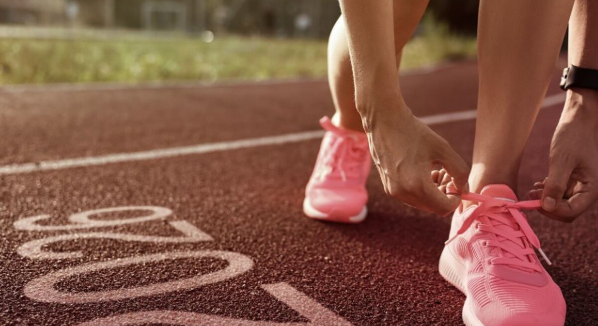 Eine Person bindet die Schnürsenkel ihrer pinken Sportschuhe, während sie auf einer Laufbahn steht. Im Vordergrund ist die Zahl "2026" auf dem Boden der Laufbahn zu sehen. Im Hintergrund sind unscharf Grünflächen und Gebäude erkennbar.