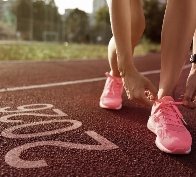 Eine Person bindet die Schnürsenkel ihrer pinken Sportschuhe, während sie auf einer Laufbahn steht. Im Vordergrund ist die Zahl "2026" auf dem Boden der Laufbahn zu sehen. Im Hintergrund sind unscharf Grünflächen und Gebäude erkennbar. | © Africa images via Canva.com
