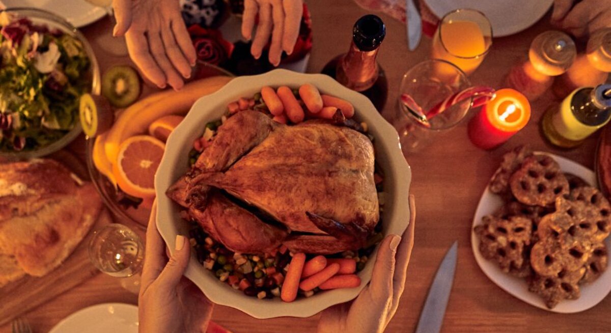 Hände reichen eine Schüssel mit gebratenem Geflügel und Gemüse über einen festlich gedeckten Tisch mit Kerzen, Getränken und Weihnachtsgebäck.