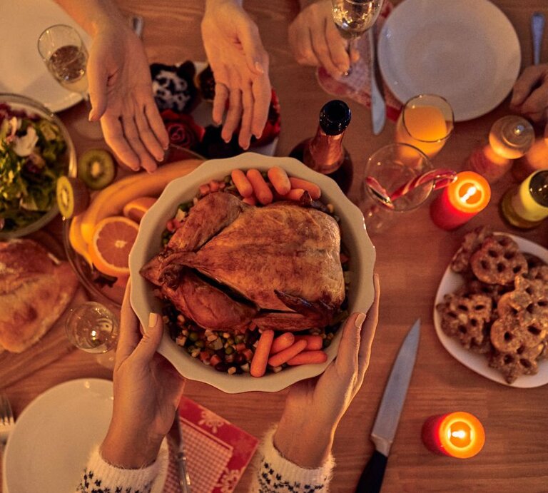 Hände reichen eine Schüssel mit gebratenem Geflügel und Gemüse über einen festlich gedeckten Tisch mit Kerzen, Getränken und Weihnachtsgebäck. | © Getty Images/ Vasyl Dolmatov via Canva.com
