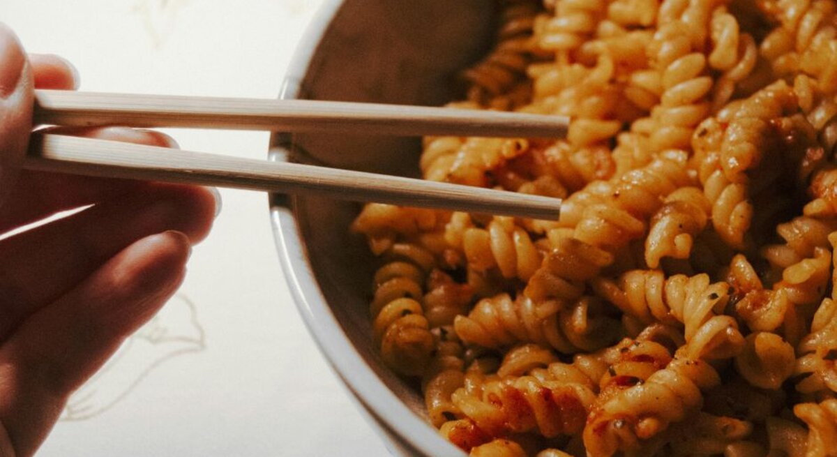 Hand hält Holzstäbchen über einer Schüssel mit Fusilli-Nudeln in rötlicher Soße auf hellem Untergrund.