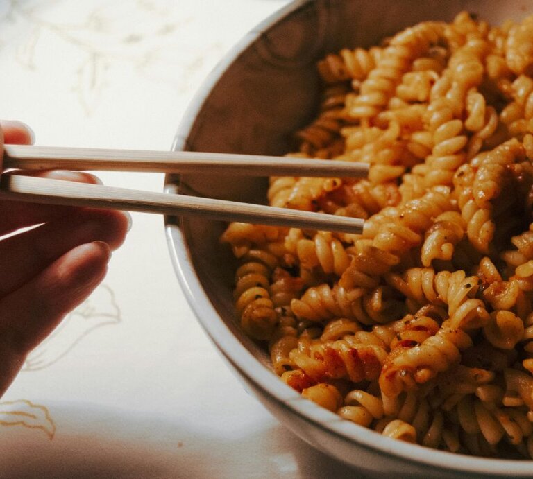 Hand hält Holzstäbchen über einer Schüssel mit Fusilli-Nudeln in rötlicher Soße auf hellem Untergrund. | © Pexels/ Esra Kaya via Canva.com 