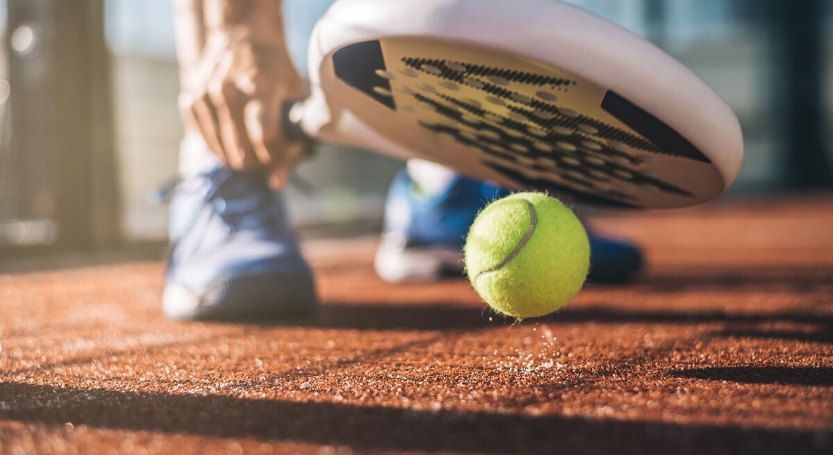 Eine Person in blauen Sportschuhen bereitet sich darauf vor, mit einem Padelschläger einen gelben Tennisball auf einem sandigen Platz zu schlagen.