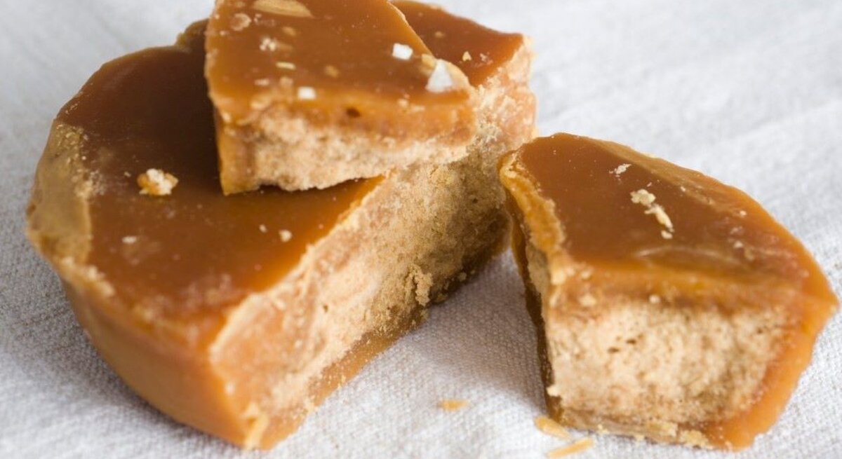 Das Bild zeigt Stücke von Jaggery, einem traditionellen, unraffinierten Zuckerprodukt, das oft in Südasien verwendet wird. Die goldbraune Färbung und die erkennbare Körnung weisen auf die natürliche Beschaffenheit hin. Der Jaggery scheint frisch gebrochen zu sein, was durch die unregelmäßigen Kanten und die sichtbaren Kristalle innerhalb des Zuckers deutlich wird. Die Textur sieht klebrig und feucht aus, typisch für hochwertigen Jaggery. Er liegt auf einer weißen Unterlage, die seine rustikale, natürliche Ästhetik betont.