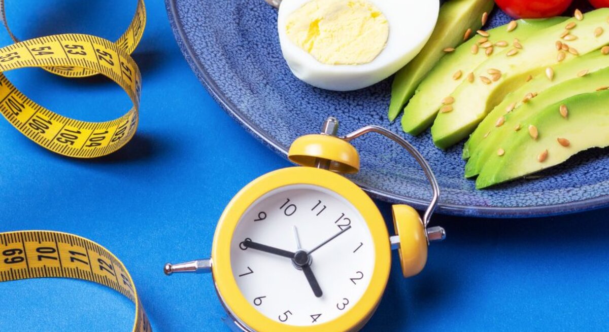 Ein Teller mit gesunden Lebensmitteln wie hartgekochten Eiern, Avocado-Scheiben mit Samen und Kirschtomaten, daneben ein gelber Wecker und ein Maßband auf einem blauen Hintergrund – Darstellung von Intervallfasten und gesunder Ernährung.