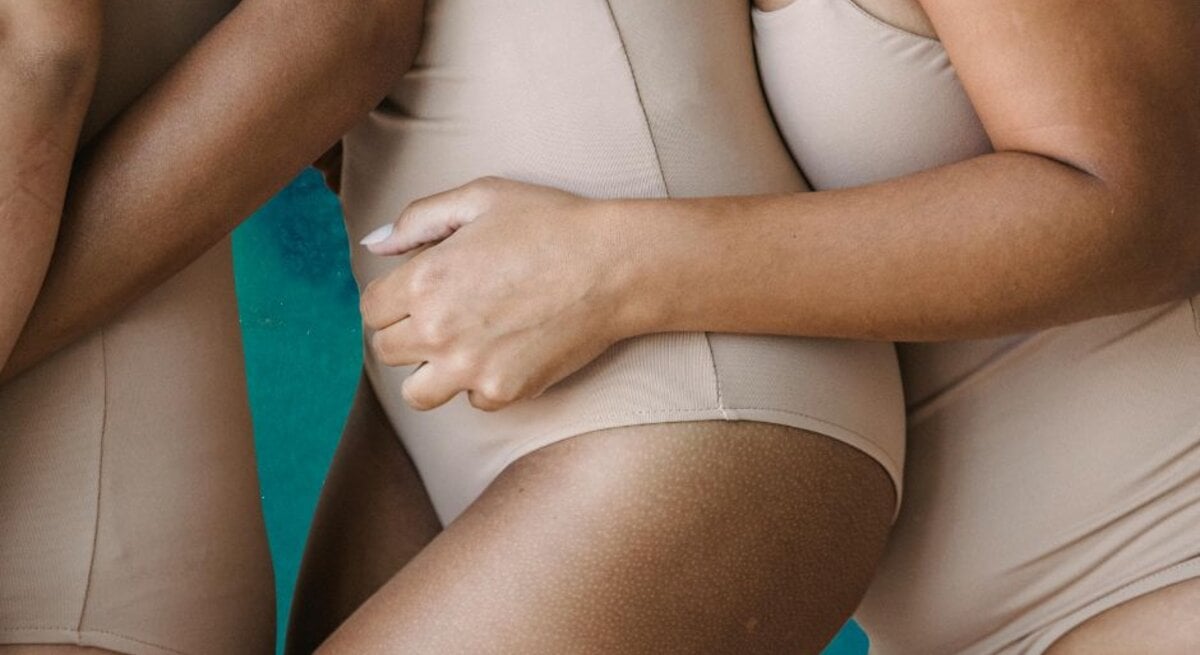 Nahaufnahme von drei Frauen in hautfarbenen Badeanzügen, die eng aneinander stehen, mit einem Hauch von blaugrünem Hintergrund.