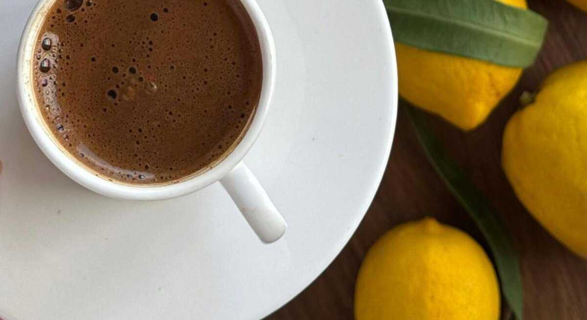Tasse mit frisch aufgebrühtem, schaumigem Kaffee auf einer weißen Untertasse, daneben mehrere gelbe Zitronen auf einer Holzoberfläche.