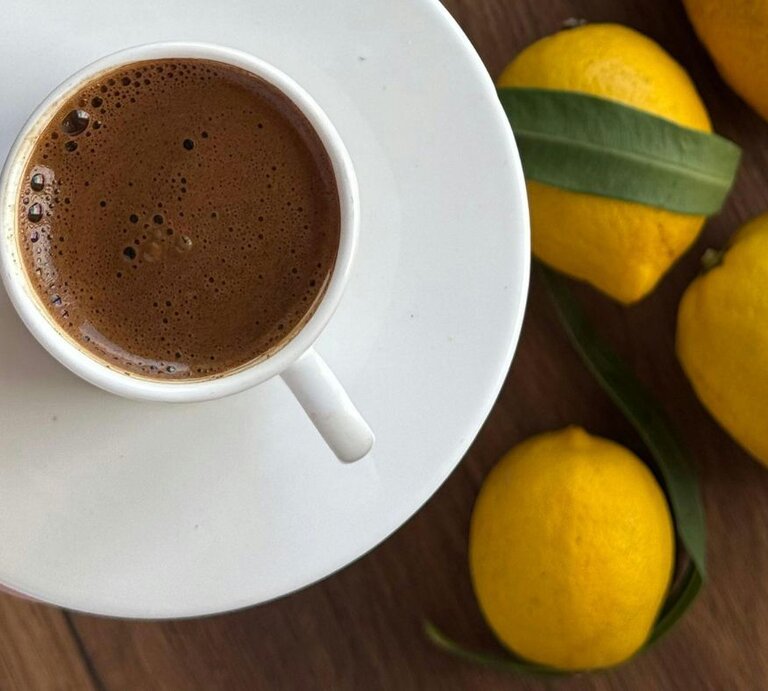 Tasse mit frisch aufgebrühtem, schaumigem Kaffee auf einer weißen Untertasse, daneben mehrere gelbe Zitronen auf einer Holzoberfläche. | © Pexels/ Merve Arlı