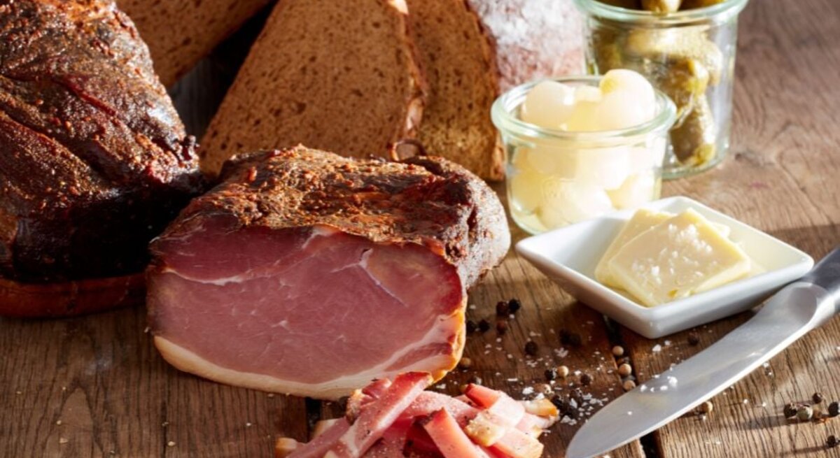 Rustikales Stillleben mit aufgeschnittenem geräuchertem Schinken auf einem Holztisch, dazu in Scheiben geschnittenes Bauernbrot, ein Messer, Gewürze sowie kleine Gläser mit Essiggurken und Perlzwiebeln und ein Tellerchen mit Butterstückchen.