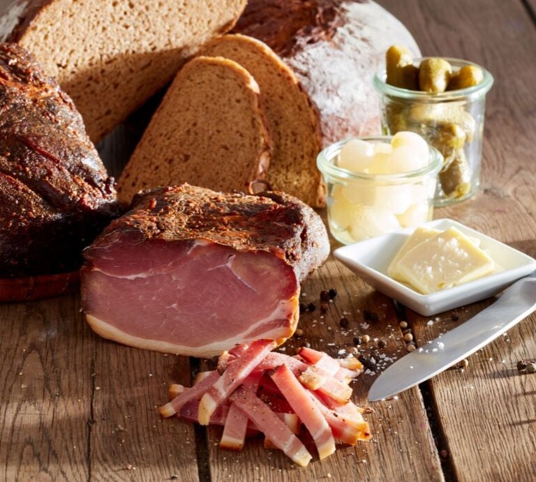 Rustikales Stillleben mit aufgeschnittenem geräuchertem Schinken auf einem Holztisch, dazu in Scheiben geschnittenes Bauernbrot, ein Messer, Gewürze sowie kleine Gläser mit Essiggurken und Perlzwiebeln und ein Tellerchen mit Butterstückchen. | © Adobe Stock/ Michael Gerlach