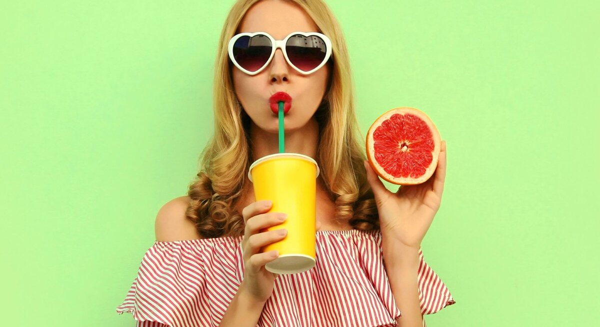 Eine blonde Frau vor grünem Hintergrund. Sie trägt eine Sonnenbrille, hält eine aufgeschnitte Grapefruit hoch und trinkt mit einem Strohhalm aus einem Becher.