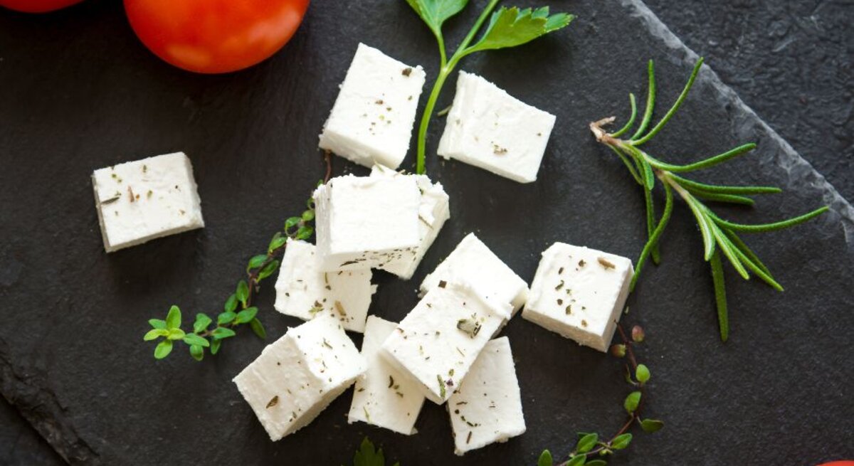 Weiße Feta-Würfel mit Kräutern auf einer dunklen Schieferplatte, umgeben von frischen Tomaten, Petersilie, Rosmarin und Thymian.