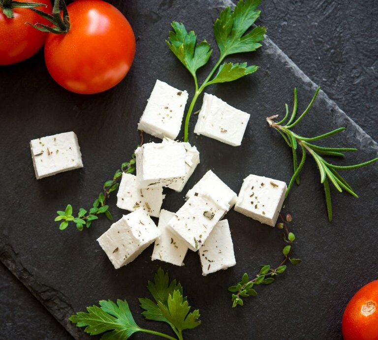 Weiße Feta-Würfel mit Kräutern auf einer dunklen Schieferplatte, umgeben von frischen Tomaten, Petersilie, Rosmarin und Thymian. | © Getty Images/ Mizina via Canva.com 