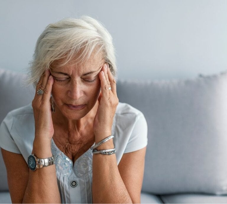 Frau mit grauen Haaren sitzt auf einem Sofa und hält sich mit beiden Händen die Schläfen. | © Getty Images Pro/ dragana991 via Canva.com 