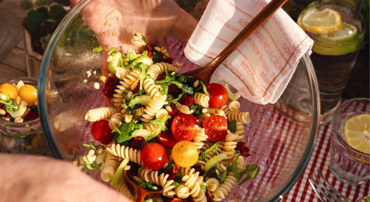 Nahaufnahme einer Glasschüssel mit mediterranem Nudelsalat (Fusilli, Rucola, rote und gelbe Kirschtomaten, Pinienkerne, getrocknete Tomaten), von einer Hand mit Holzbesteck gemischt; Picknick auf rot-weißem Tischtuch, Zitronenwasser im Hintergrund.