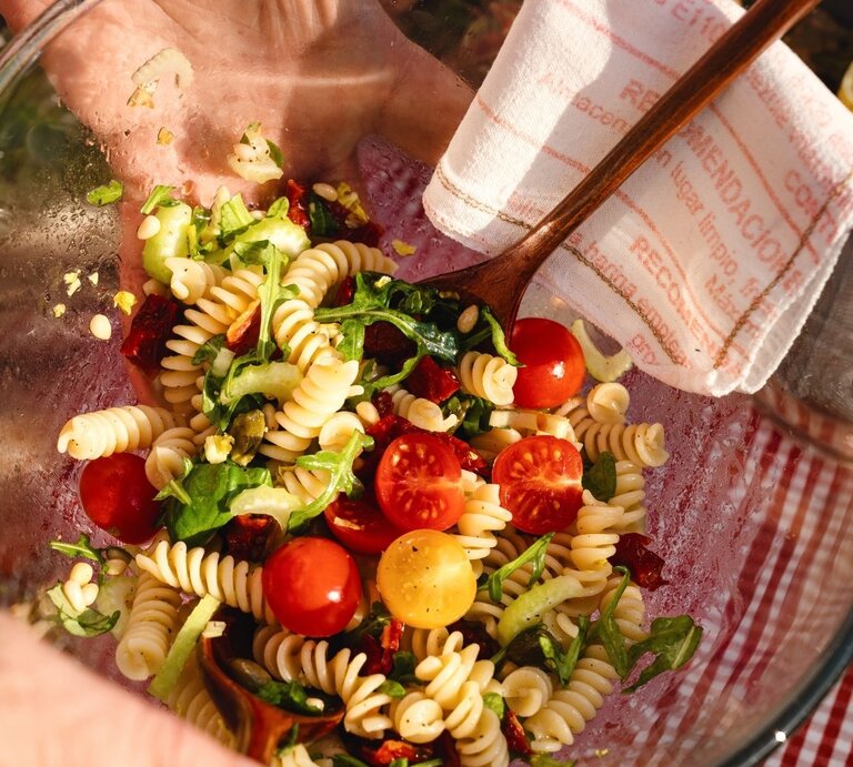 Nahaufnahme einer Glasschüssel mit mediterranem Nudelsalat (Fusilli, Rucola, rote und gelbe Kirschtomaten, Pinienkerne, getrocknete Tomaten), von einer Hand mit Holzbesteck gemischt; Picknick auf rot-weißem Tischtuch, Zitronenwasser im Hintergrund. | © Adobe Stock/ MC Stock