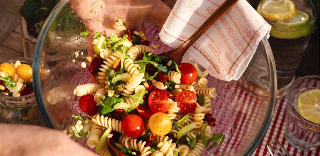 Nahaufnahme einer Glasschüssel mit mediterranem Nudelsalat (Fusilli, Rucola, rote und gelbe Kirschtomaten, Pinienkerne, getrocknete Tomaten), von einer Hand mit Holzbesteck gemischt; Picknick auf rot-weißem Tischtuch, Zitronenwasser im Hintergrund. | © Adobe Stock/ MC Stock