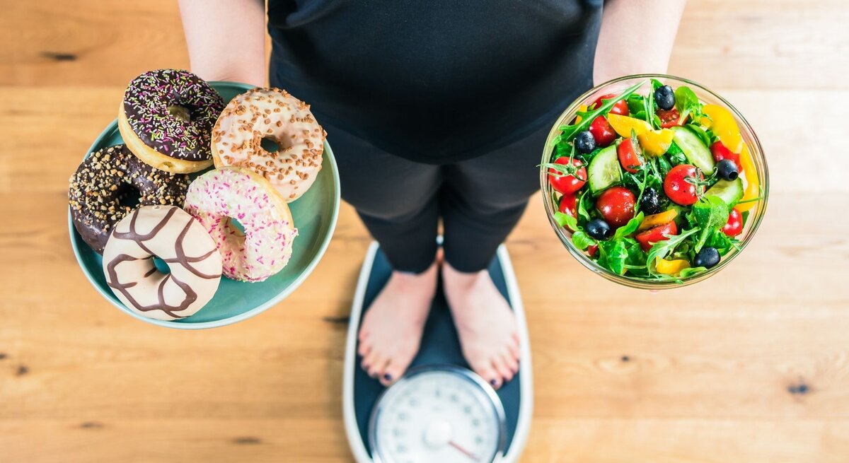 Eine Person steht auf einer Waage und hält zwei Schalen: links eine mit fünf bunten Donuts, rechts eine mit frischem Salat aus Gemüse und Beeren. Das Bild zeigt den Gegensatz zwischen ungesunden Süßigkeiten und gesunder Ernährung.