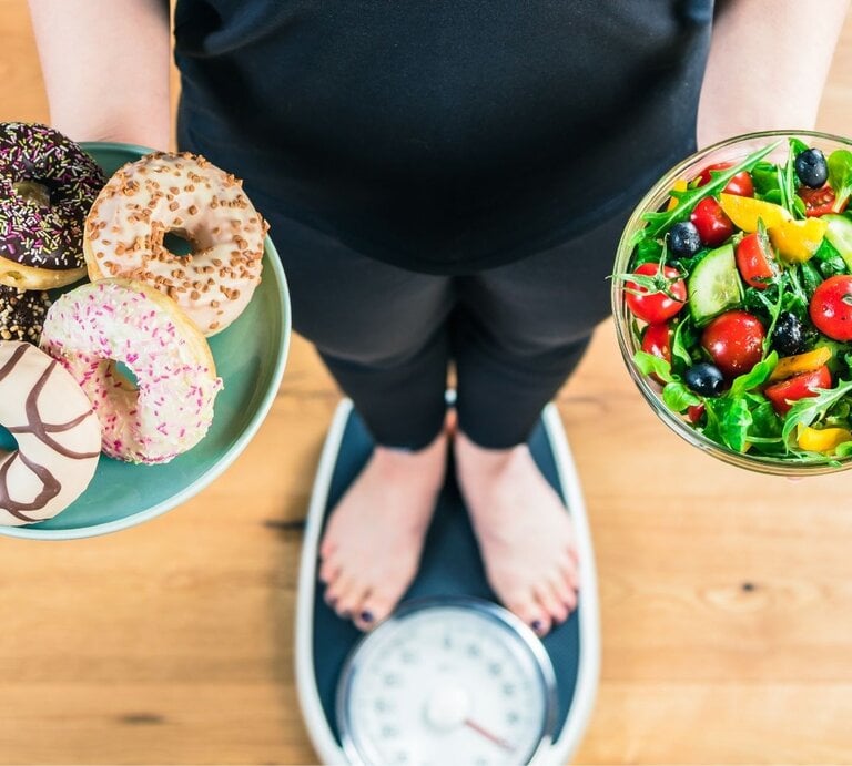 Eine Person steht auf einer Waage und hält zwei Schalen: links eine mit fünf bunten Donuts, rechts eine mit frischem Salat aus Gemüse und Beeren. Das Bild zeigt den Gegensatz zwischen ungesunden Süßigkeiten und gesunder Ernährung. | © Adobe Stock/ karepa