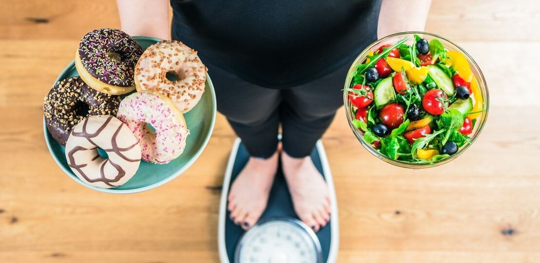 Eine Person steht auf einer Waage und hält zwei Schalen: links eine mit fünf bunten Donuts, rechts eine mit frischem Salat aus Gemüse und Beeren. Das Bild zeigt den Gegensatz zwischen ungesunden Süßigkeiten und gesunder Ernährung. | © Adobe Stock/ karepa