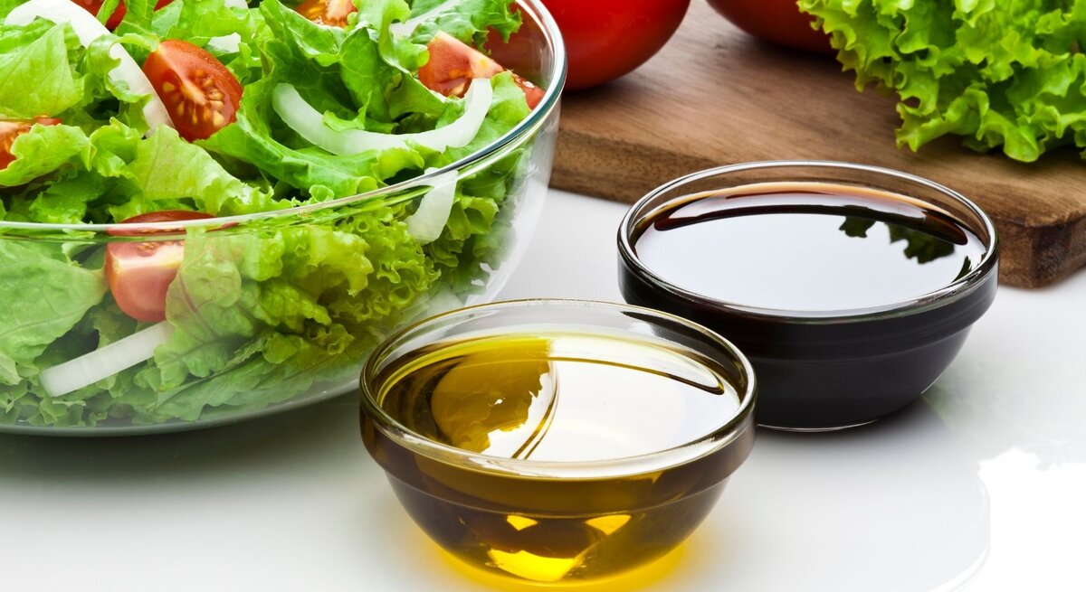Glasschüssel mit grünem Salat, Tomaten und Zwiebeln; daneben zwei Schälchen mit Olivenöl und dunklem Balsamico.