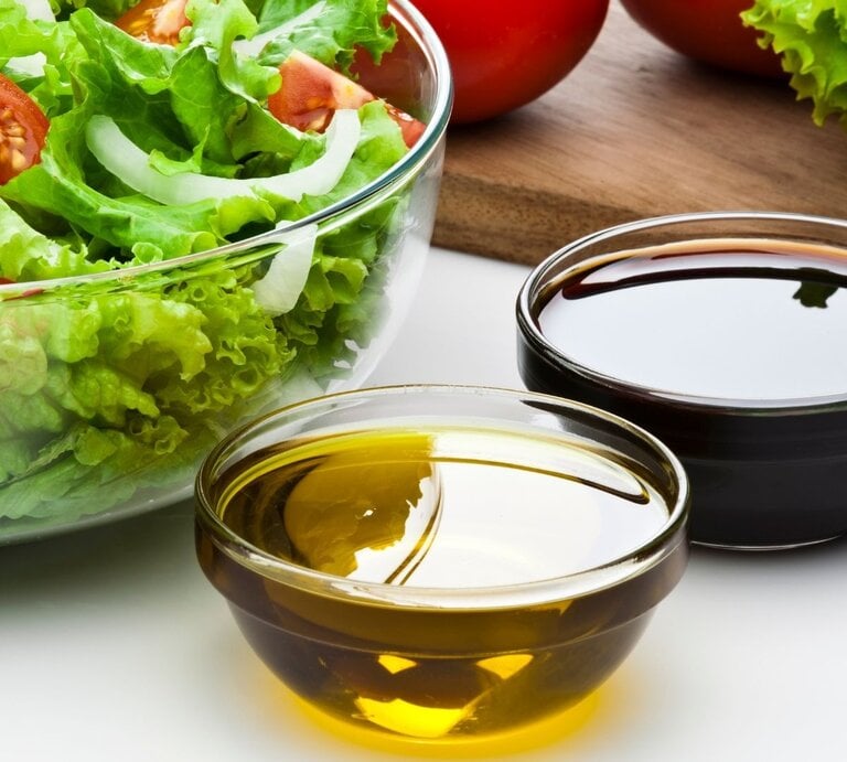 Glasschüssel mit grünem Salat, Tomaten und Zwiebeln; daneben zwei Schälchen mit Olivenöl und dunklem Balsamico. | © Getty Images Signature/ fcafotodigital via Canva.com
