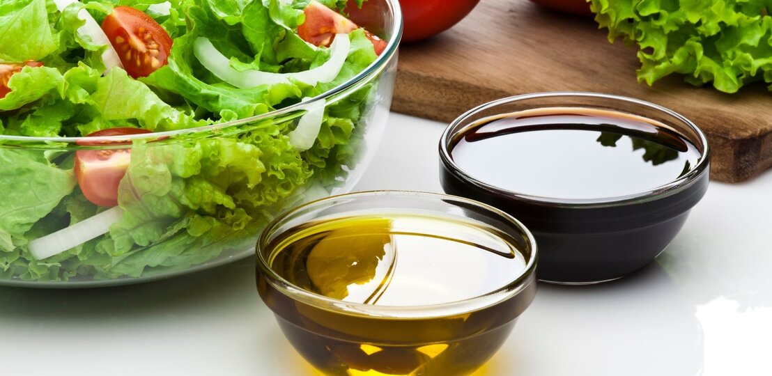 Glasschüssel mit grünem Salat, Tomaten und Zwiebeln; daneben zwei Schälchen mit Olivenöl und dunklem Balsamico. | © Getty Images Signature/ fcafotodigital via Canva.com