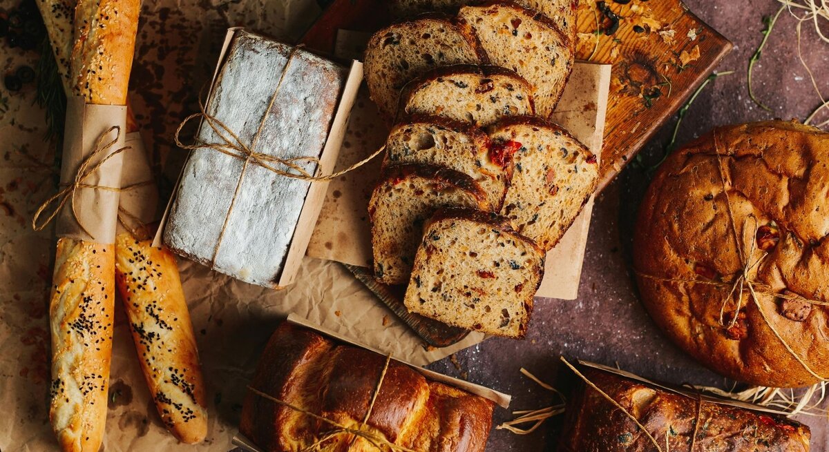 Verschiedene Brotsorten liegen dekorativ auf braunem Untergrund. Zu sehen sind Baguettes mit Sesam, ein mit Mehl bestäubtes Kastenbrot, geschnittenes Körnerbrot mit Tomatenstücken und ein rundes, rustikales Brot. Einige Brote sind mit Papier und Kordel umwickelt, im Hintergrund liegen Kräuter und Oliven.