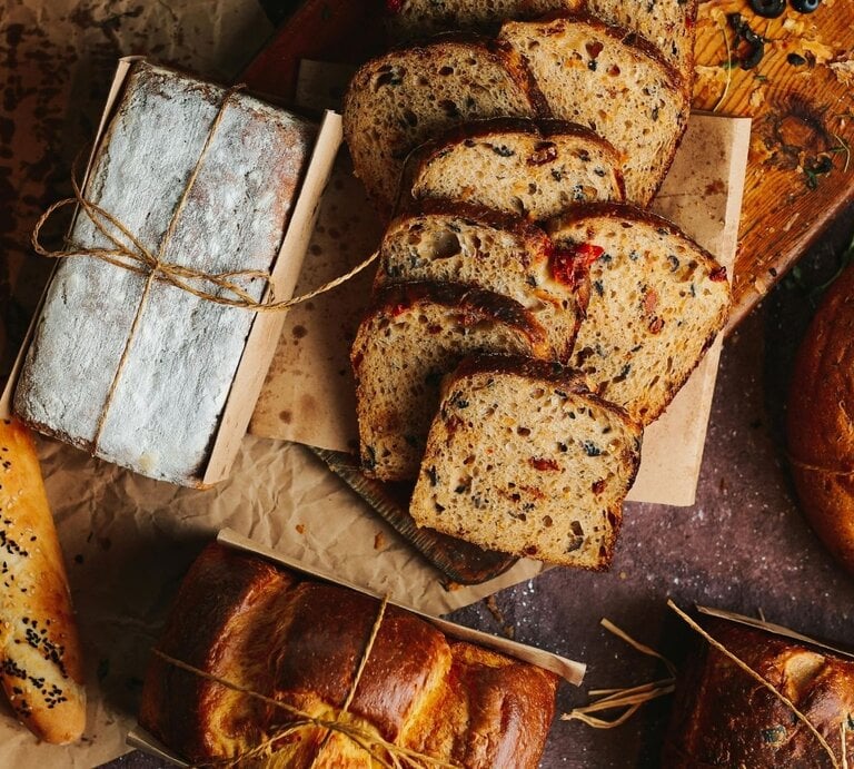 Verschiedene Brotsorten liegen dekorativ auf braunem Untergrund. Zu sehen sind Baguettes mit Sesam, ein mit Mehl bestäubtes Kastenbrot, geschnittenes Körnerbrot mit Tomatenstücken und ein rundes, rustikales Brot. Einige Brote sind mit Papier und Kordel umwickelt, im Hintergrund liegen Kräuter und Oliven. | © Pexels/ Taha Samet Arslan via Canva.com 