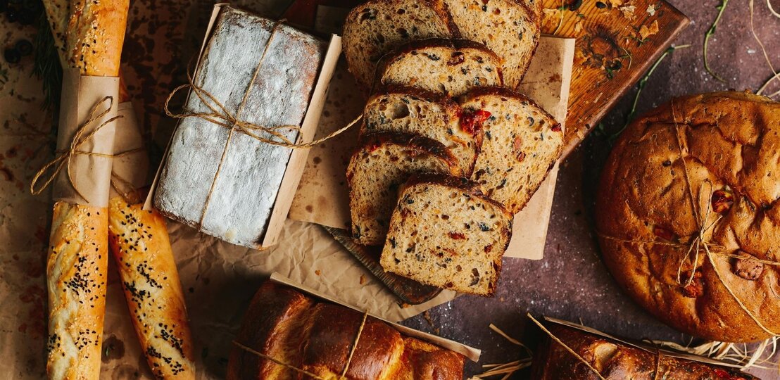 Verschiedene Brotsorten liegen dekorativ auf braunem Untergrund. Zu sehen sind Baguettes mit Sesam, ein mit Mehl bestäubtes Kastenbrot, geschnittenes Körnerbrot mit Tomatenstücken und ein rundes, rustikales Brot. Einige Brote sind mit Papier und Kordel umwickelt, im Hintergrund liegen Kräuter und Oliven. | © Pexels/ Taha Samet Arslan via Canva.com 