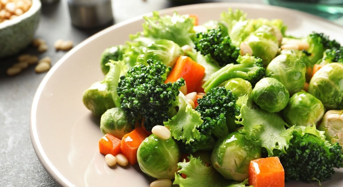 Nahaufnahme eines frischen Gemüsetellers mit Brokkoli, Rosenkohl, Blattsalat, Karottenwürfeln und Pinienkernen in einer hellen Schüssel.