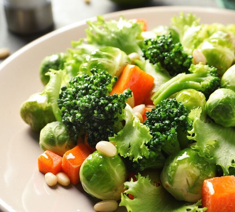 Nahaufnahme eines frischen Gemüsetellers mit Brokkoli, Rosenkohl, Blattsalat, Karottenwürfeln und Pinienkernen in einer hellen Schüssel. | © Africa images via Canva.com