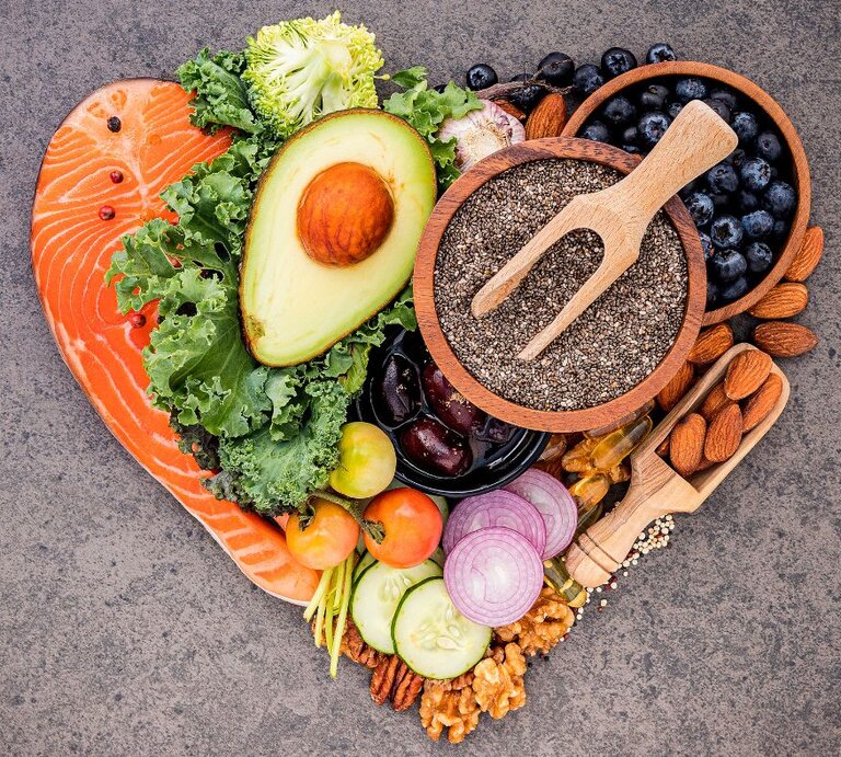 Ein herzförmiges Arrangement aus gesunden Lebensmitteln, darunter Lachs, Avocado, Grünkohl, Chiasamen, Blaubeeren, Mandeln, Walnüsse, Kirschtomaten, rote Zwiebeln, Gurkenscheiben, Brokkoli und Knoblauch. Die Zutaten sind farbenfroh präsentiert und symbolisieren eine ausgewogene Ernährung.   Fragen  Erklären | © Canva.com