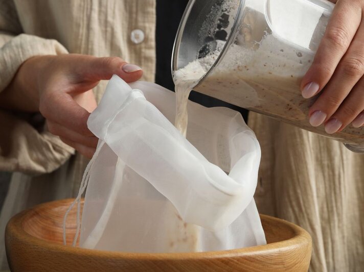 Eine Person gießt selbstgemachte Mandelmilch aus einem Mixer durch einen feinmaschigen Stoffbeutel in eine Holzschale, um die Feststoffe vom Flüssigen zu trennen. Die Hände halten den Beutel und den Mixer, im Hintergrund ist Kleidung zu erkennen. | © Canva.com