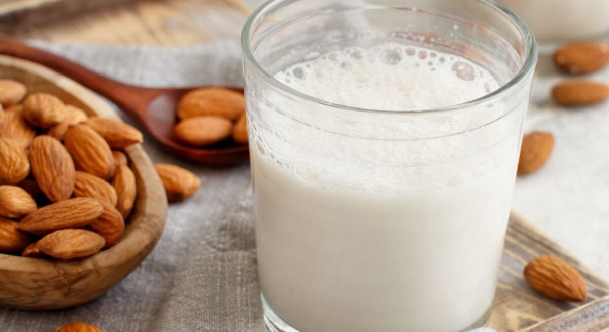 Ein Glas frisch zubereitete Mandelmilch steht auf einem Holzbrett, umgeben von ganzen Mandeln in einer Holzschale sowie einer Stoffserviette. Im Hintergrund ist ein weiteres Glas mit Mandelmilch unscharf zu erkennen.