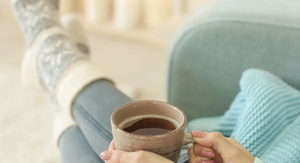 Frau in gemütlicher Kleidung sitzt entspannt auf einem Sofa, trägt dicke Stricksocken und hält eine Tasse Tee oder Kaffee in den Händen, im unscharfen Hintergrund warme Kerzen- und Lichterdekoration.