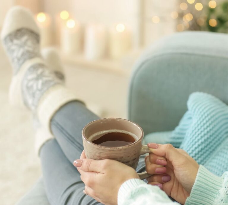 Frau in gemütlicher Kleidung sitzt entspannt auf einem Sofa, trägt dicke Stricksocken und hält eine Tasse Tee oder Kaffee in den Händen, im unscharfen Hintergrund warme Kerzen- und Lichterdekoration. | © pixelshot via Canva.com
