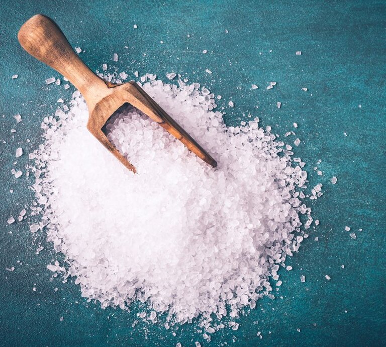 Ein Holzlöffel liegt auf grobem Meersalz, das auf einer grünlichen Unterlage verstreut ist. | © Adobe Stock/ Fischer Food Design