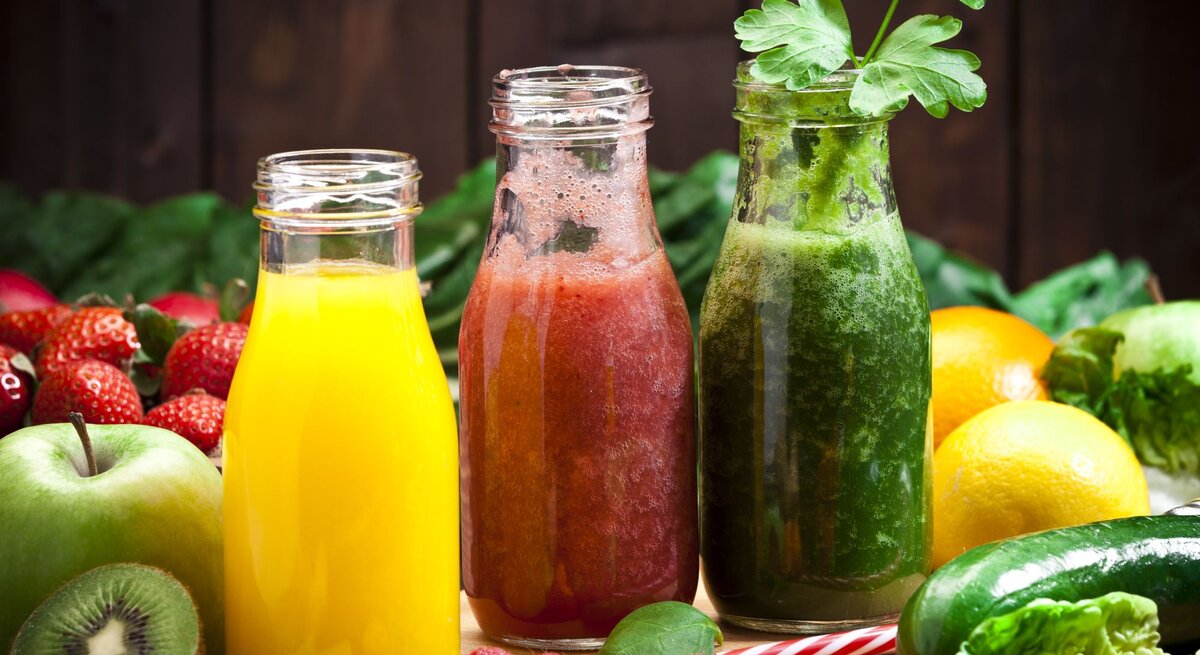 Es sind drei Glasflaschen zu sehen, die mit bunten Säften für eine Saftkur gefüllt sind. Im Hintergrund liegt weiteres Obst und Gemüse.