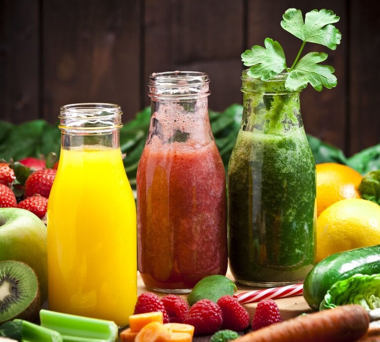 Es sind drei Glasflaschen zu sehen, die mit bunten Säften für eine Saftkur gefüllt sind. Im Hintergrund liegt weiteres Obst und Gemüse. | © gettyimages/ fcafotodigital