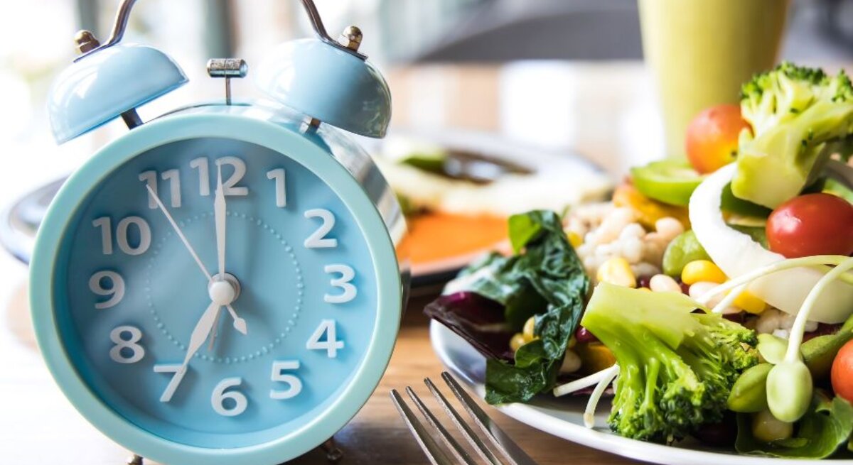 Eine hellblaue Weckeruhr auf einem Holztisch neben einem Teller mit frischem Salat. Der Salat ist bunt und beinhaltet Gemüse wie Brokkoli, Tomaten, Mais und grüne Bohnen. Im Hintergrund ist verschwommen eine weitere Mahlzeit und ein Getränk zu sehen.