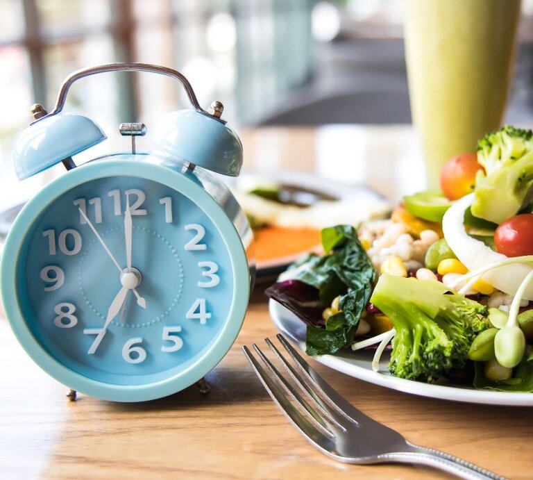 Eine hellblaue Weckeruhr auf einem Holztisch neben einem Teller mit frischem Salat. Der Salat ist bunt und beinhaltet Gemüse wie Brokkoli, Tomaten, Mais und grüne Bohnen. Im Hintergrund ist verschwommen eine weitere Mahlzeit und ein Getränk zu sehen. | © Getty Images/ Sasithorn Phuapankasemsuk via Canva.com
