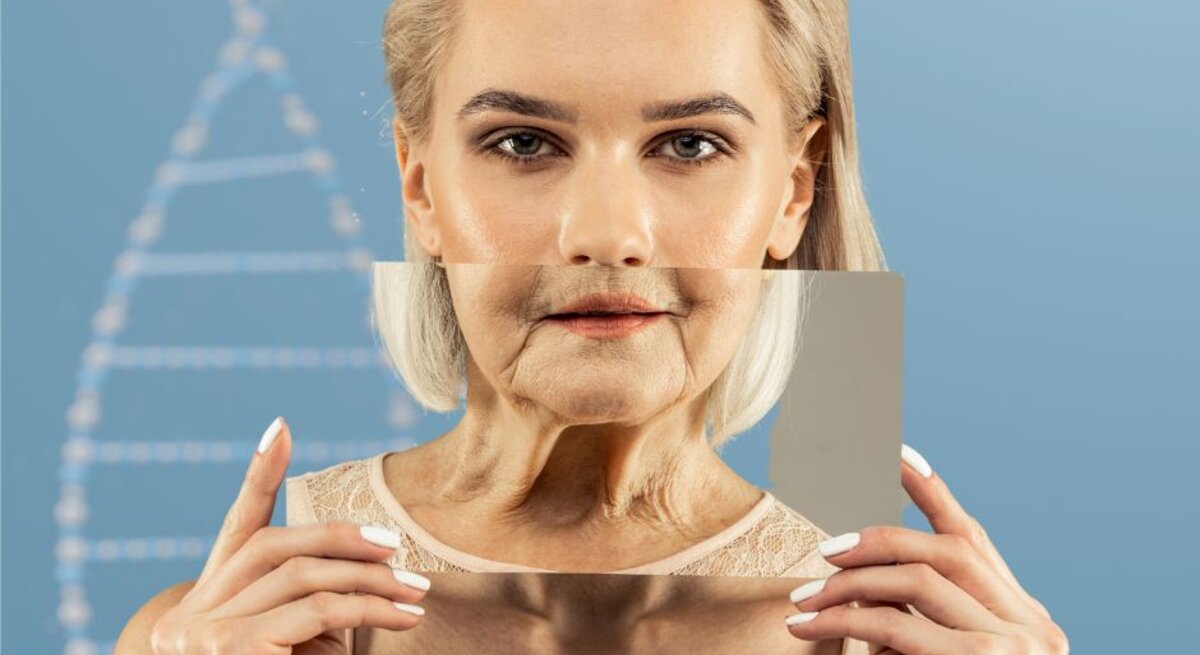 Eine Frau mit blondem Haar hält einen rechteckigen Spiegel vor ihr Gesicht. Der Spiegel zeigt ihr gealtertes Spiegelbild mit Falten und schlaffer Haut, während ihr tatsächliches Gesicht jugendlich und glatt wirkt. Im Hintergrund ist schemenhaft eine DNA-Doppelhelix zu sehen.