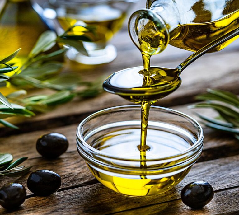 Eine Schale mit goldgelbem Olivenöl steht auf einem Holztisch; darüber gießt jemand frisches Olivenöl aus einer Flasche auf einen Löffel. Rundherum liegen Oliven und Olivenzweige. | © Getty Images Signature/ fcafotodigital via Canva.com