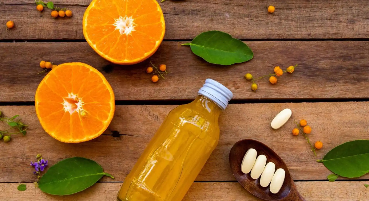 Auf einem Holztisch liegen halbierte Orangen, ein Fläschchen mit Orangensaft und ein Löffel mit Vitamin-C-Tabletten – Symbolbild für hochdosiertes Vitamin C als Nahrungsergänzung im Vergleich zur natürlichen Vitamin-C-Quelle aus Obst.